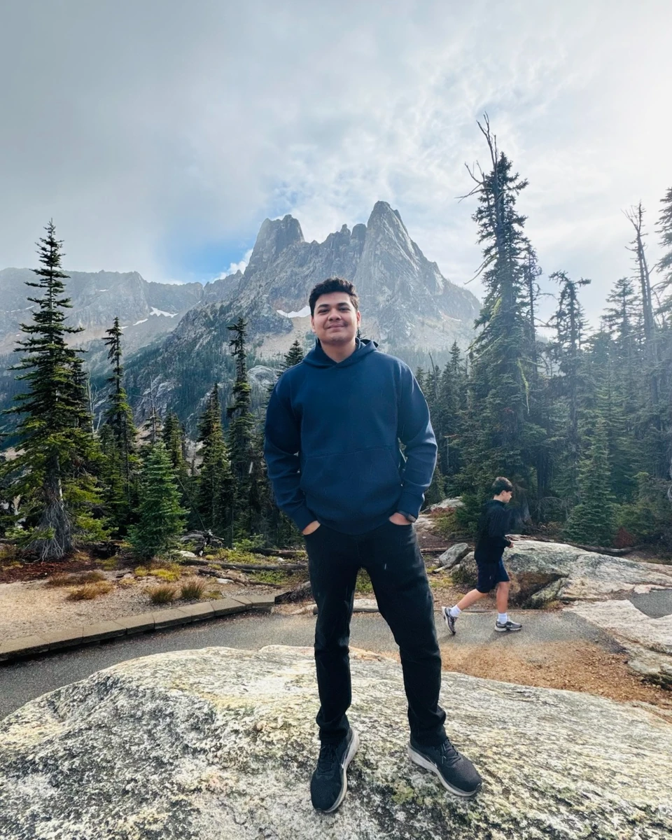 Prakhar Jain standing on a rocky overlook with alpine peaks and evergreen trees behind him.